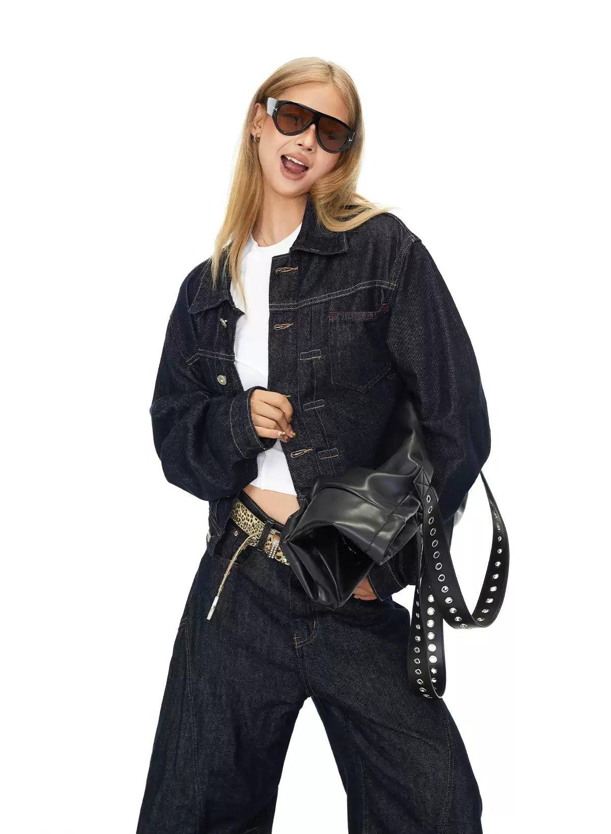 A woman wearing a denim jacket, sunglasses, and jeans is posing for a photo. She is holding a black bag with silver studs.