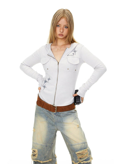 A young woman with blonde hair is standing with her hands on her hips, wearing a white long-sleeved shirt with a zipper and a brown belt. She is also wearing distressed jeans.
