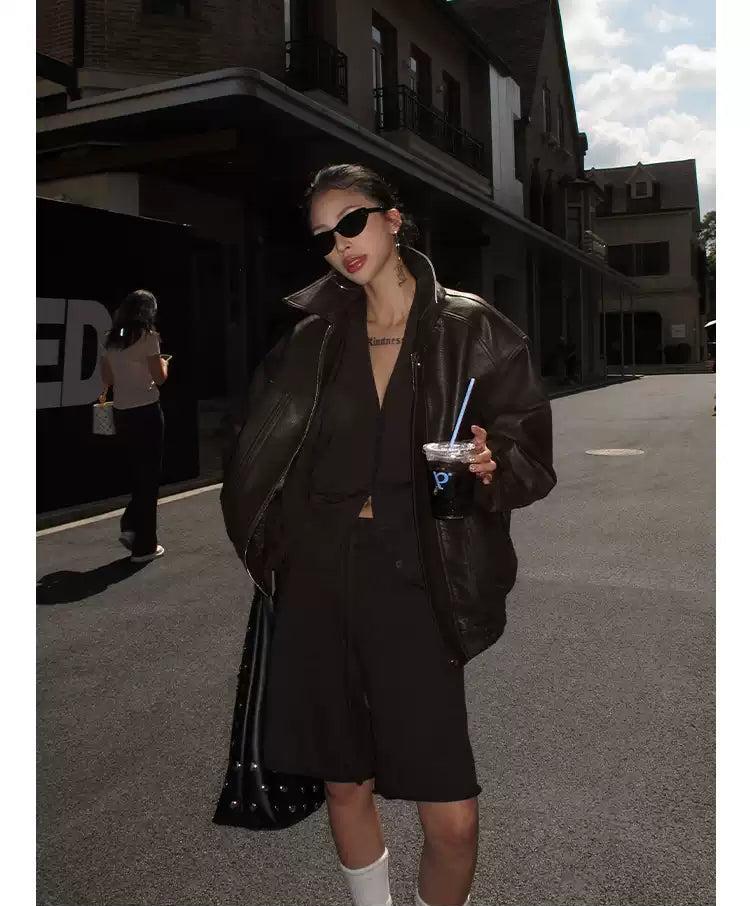 A woman wearing a black leather jacket, sunglasses, and carrying a drink stands on a street.
