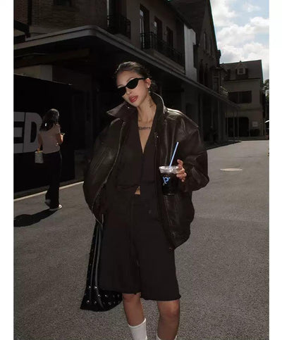 A woman wearing a black leather jacket, sunglasses, and carrying a drink stands on a street.