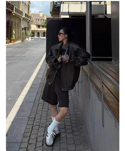 A woman in a black jacket and shorts stands on a city street, leaning against a railing and holding a drink.