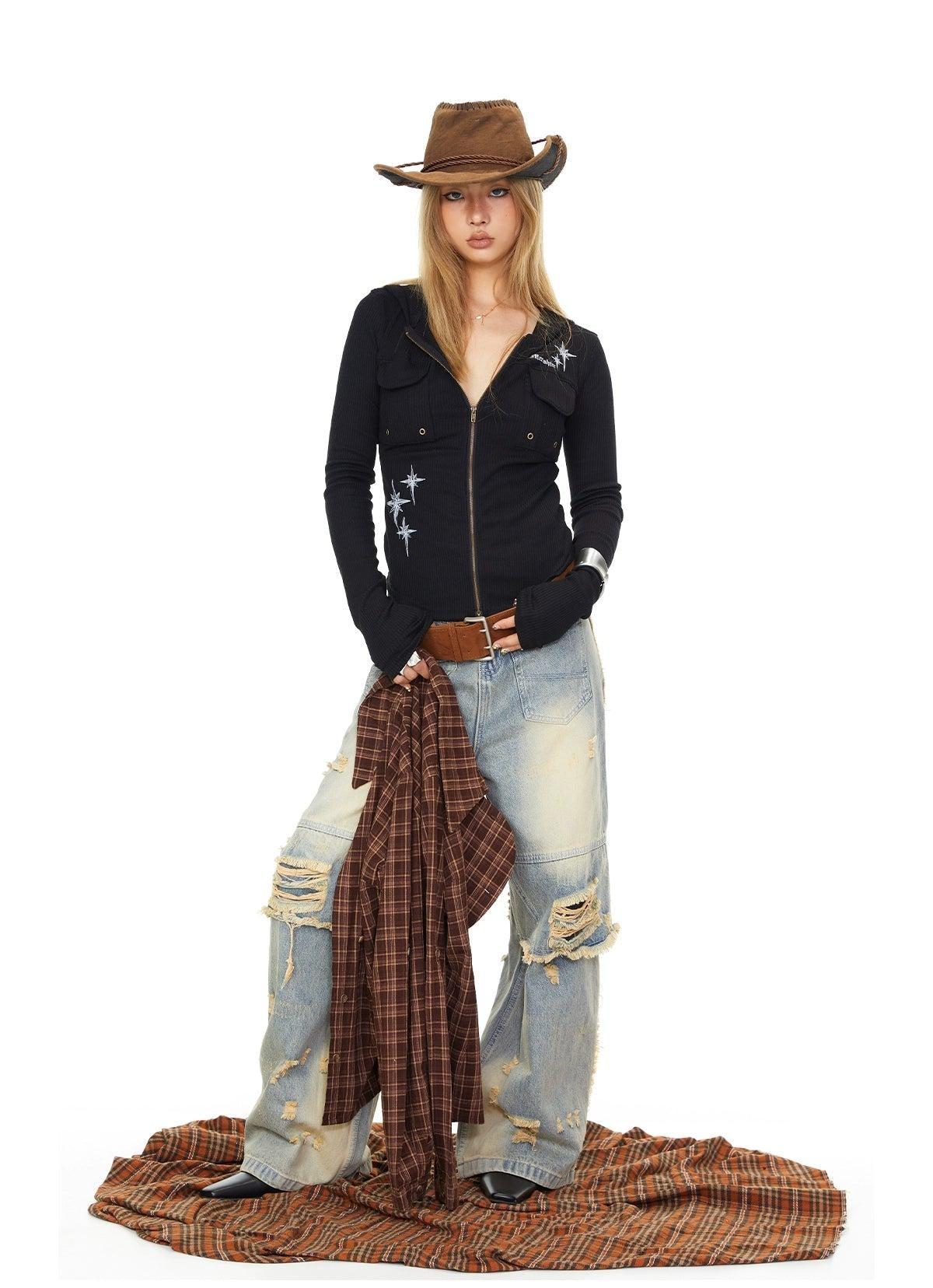 A woman wearing a black jacket, jeans, and a brown cowboy hat stands in front of a white background, holding a plaid shirt.