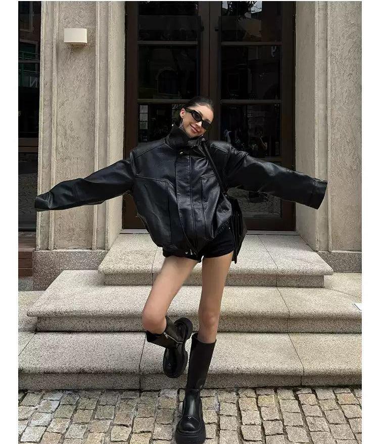 A woman wearing a black leather jacket, sunglasses, and boots is posing on a cobblestone street in front of a building.