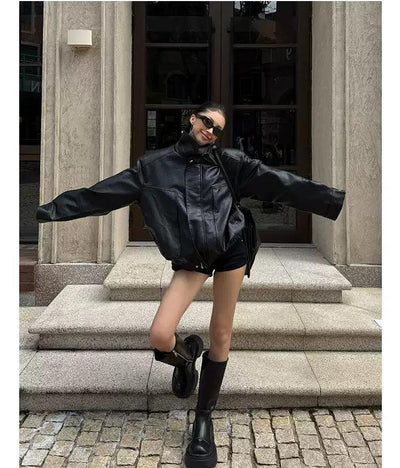 A woman wearing a black leather jacket, sunglasses, and boots is posing on a cobblestone street in front of a building.