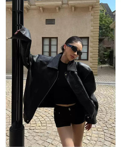 A woman wearing a black leather jacket and sunglasses stands in front of a building, leaning against a black pole.